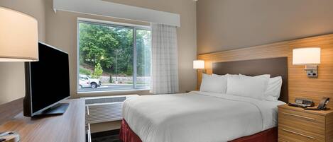 Een hotelkamer met een groot bed, een bureau met een flatscreen-televisie, een nachtkastje met een lamp en uitzicht op het omliggende landschap door een raam.