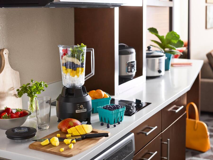Una encimera de cocina con una licuadora, una tabla de cortar con fruta, un tazón de fresas, un vaso y una planta.