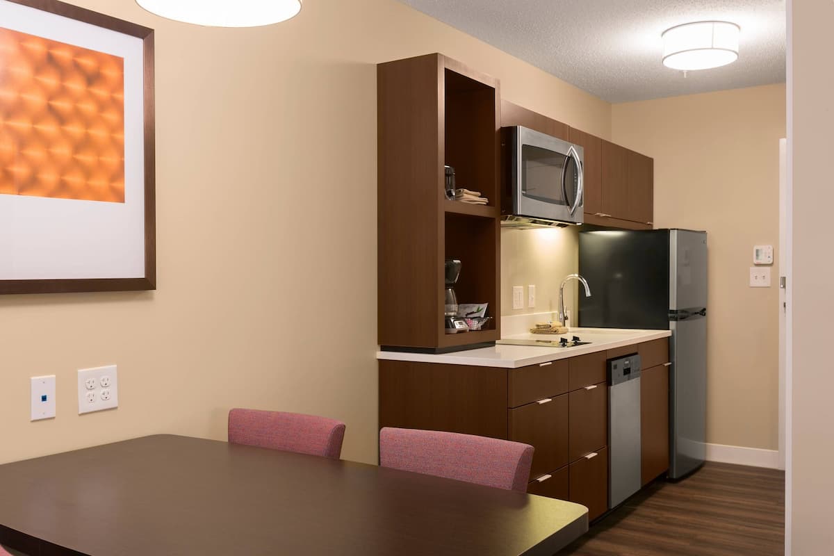 A compact kitchen with a microwave, refrigerator, and built-in dishwasher. There is a dining table with chairs and a wall-mounted light fixture.