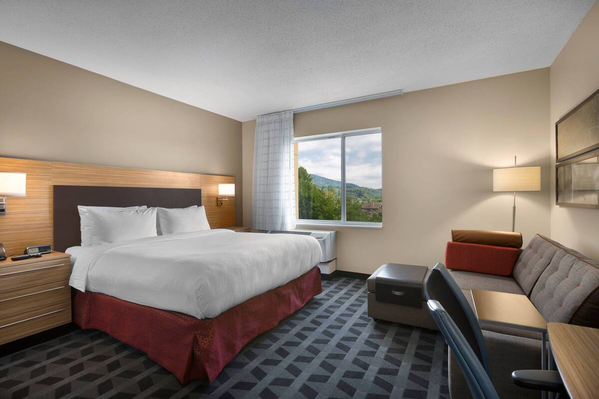 A hotel room with a large bed, a desk, a chair, a sofa, and a window with a mountain view.