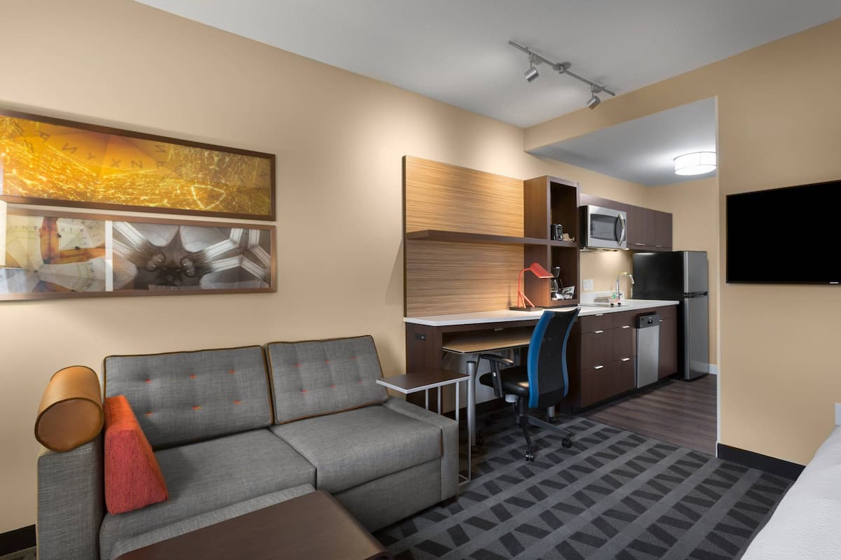 A compact hotel room with a sofa, a desk, a kitchenette, and a flat-screen TV.