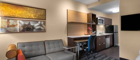 A compact hotel room with a sofa, a desk, a kitchenette, and a flat-screen TV.