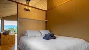 A neatly made bed with white linens and two rolled towels, a wooden bench with cushions, and a view of the outdoors through a large window.