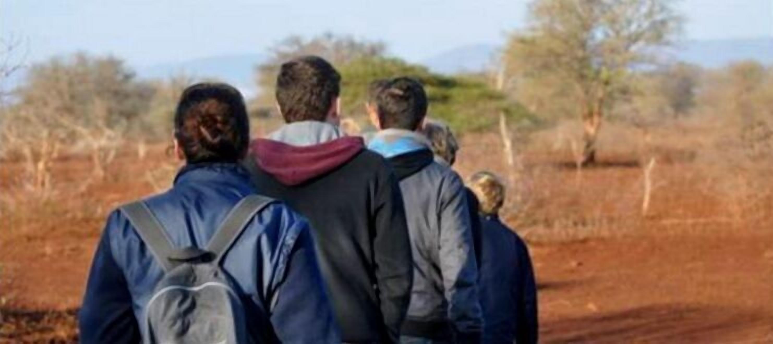 Tres personas caminando por un sendero de tierra con vegetación escasa y árboles al fondo.