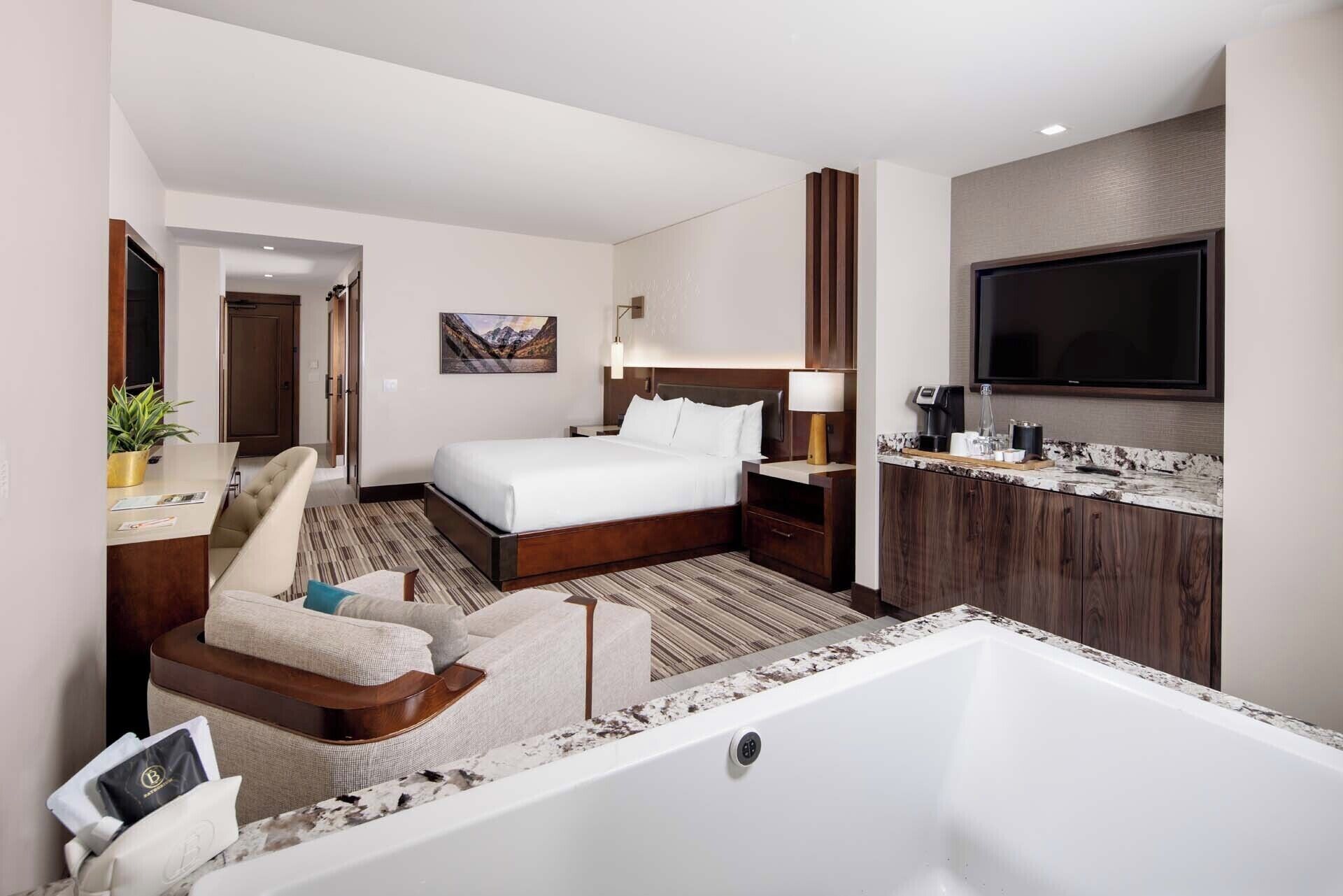 Habitación de hotel con una cama grande, un rincón de descanso con una silla y una mesita, un baño con bañera, y un televisor de pantalla plana montado en la pared.