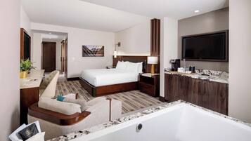 Habitación de hotel con una cama grande, un rincón de descanso con una silla y una mesita, un baño con bañera, y un televisor de pantalla plana montado en la pared.