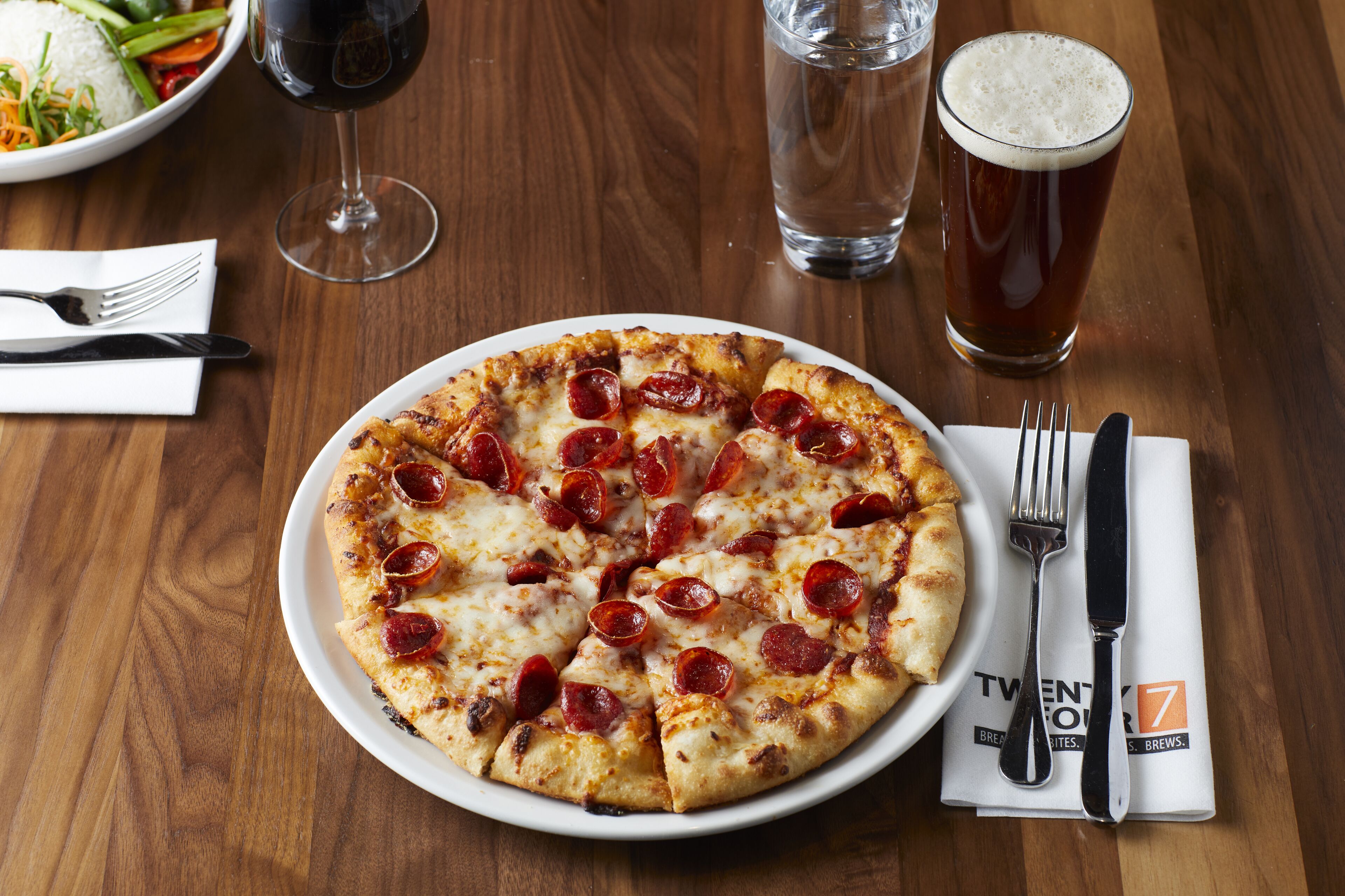 Una pizza con rodajas de pepperoni sobre un plato blanco, acompañada de una jarra de cerveza y un vaso de agua sobre una mesa de madera.