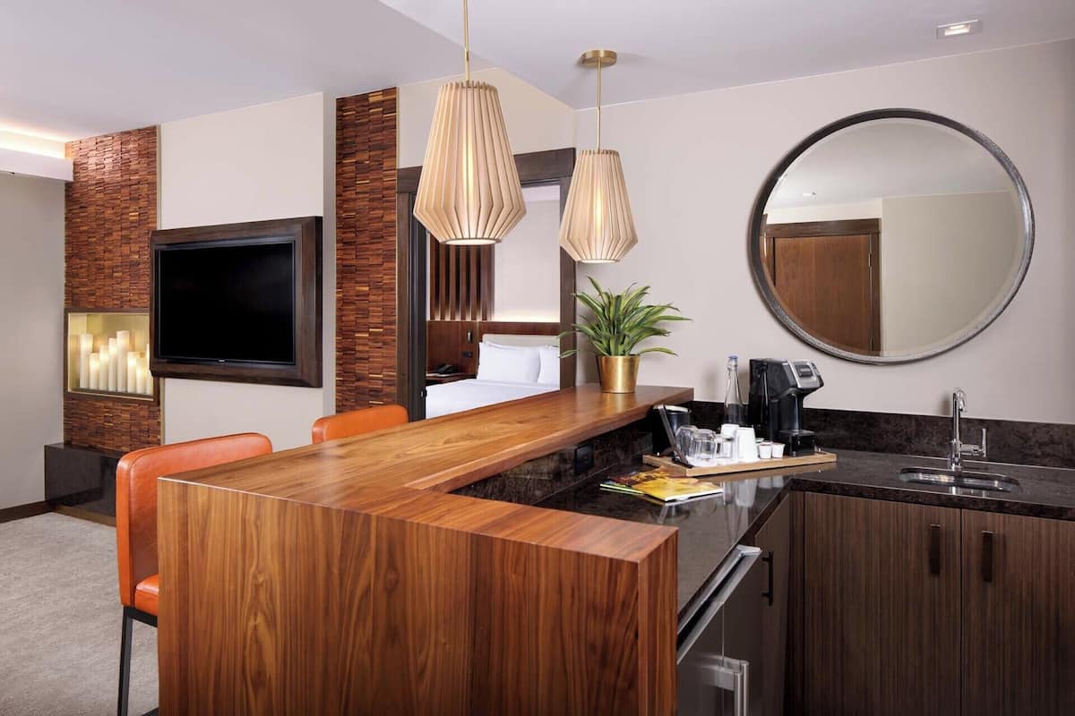 Une chambre d’hôtel moderne avec une table à manger en bois, des chaises orange, un téléviseur à écran plat, un grand miroir et un coin café.