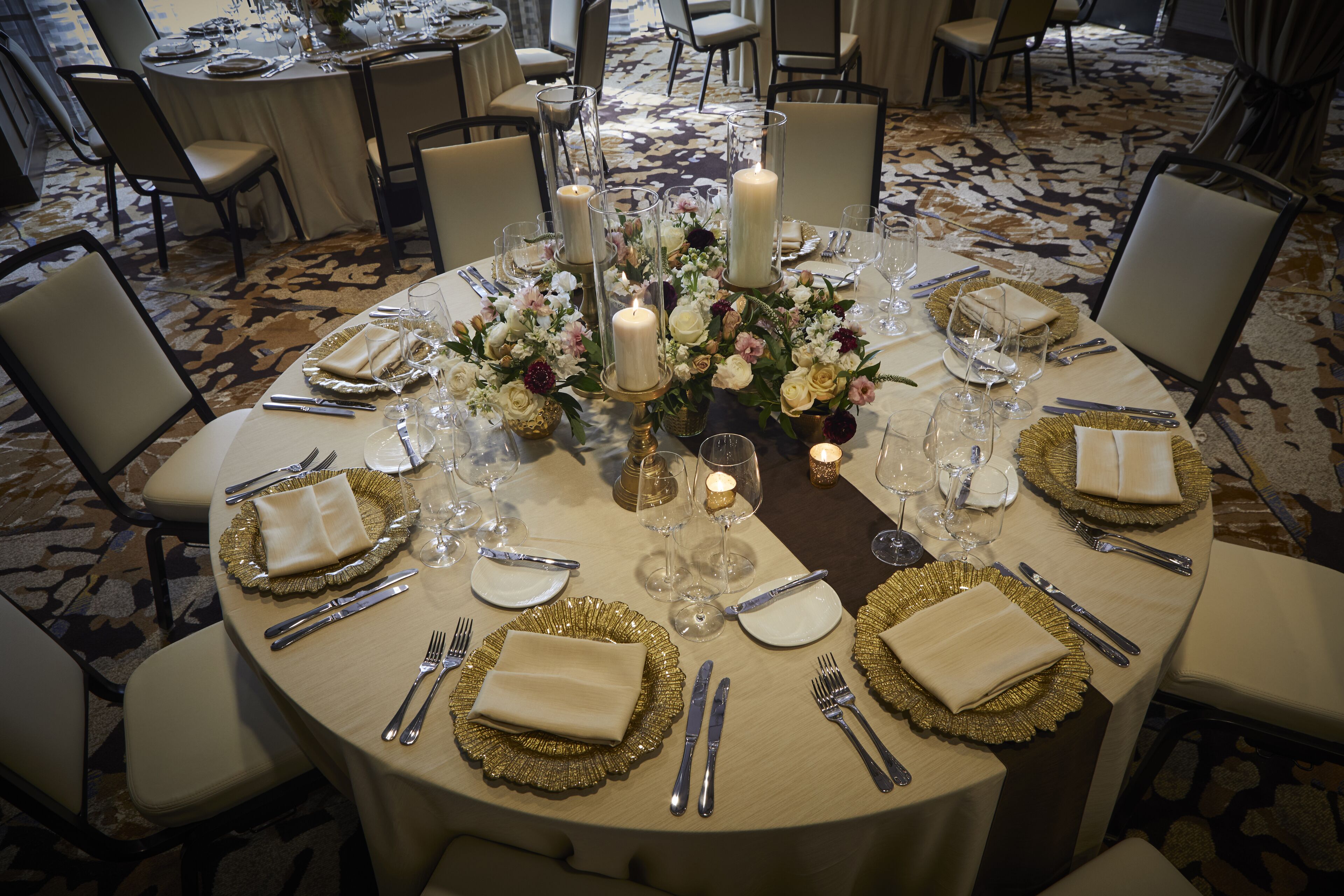 A round dining table set for a formal event with gold-rimmed plates, silverware, and candles.