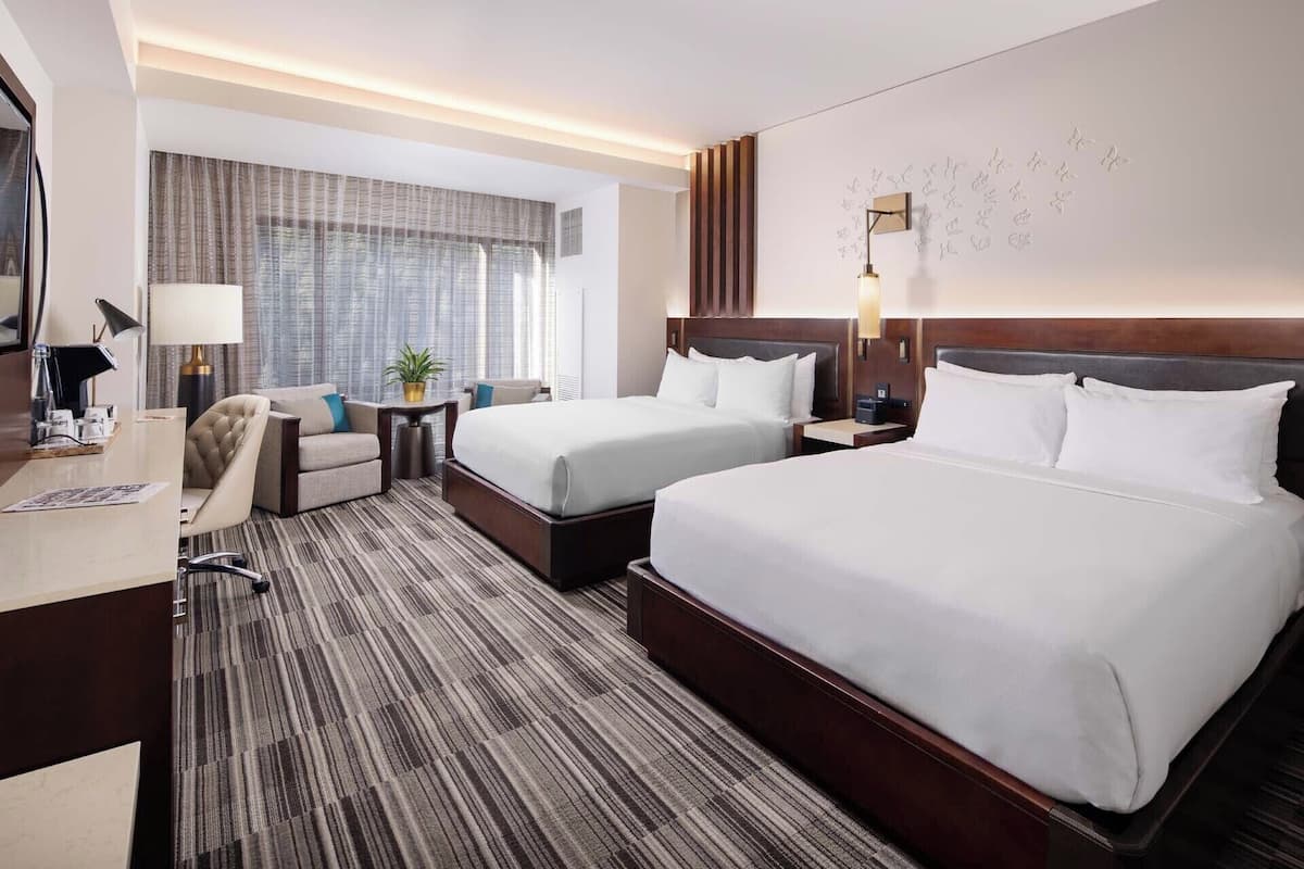 Une chambre d’hôtel avec deux lits, un bureau, une chaise, une lampe et une fenêtre avec des rideaux.