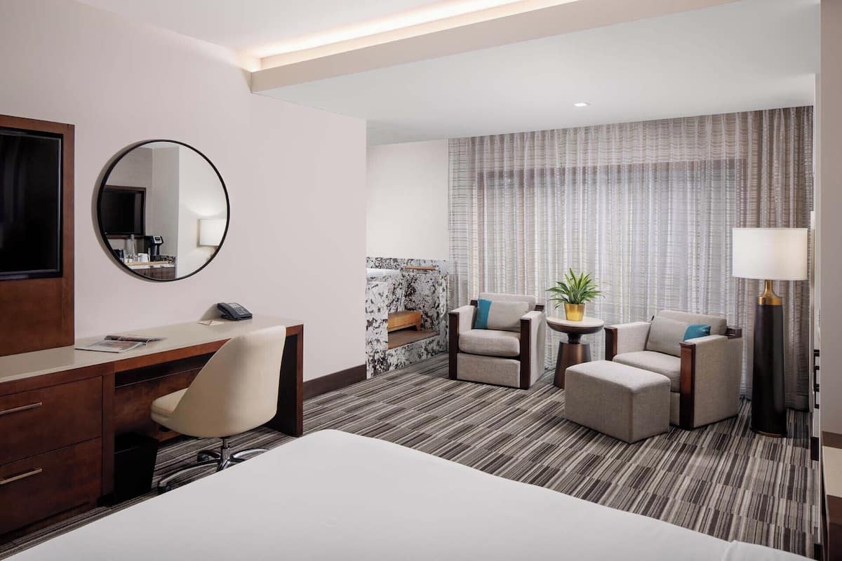 Une chambre d’hôtel comprenant un lit, un bureau, une chaise, deux fauteuils, une petite table avec une plante, un miroir rond et une grande fenêtre avec des rideaux.