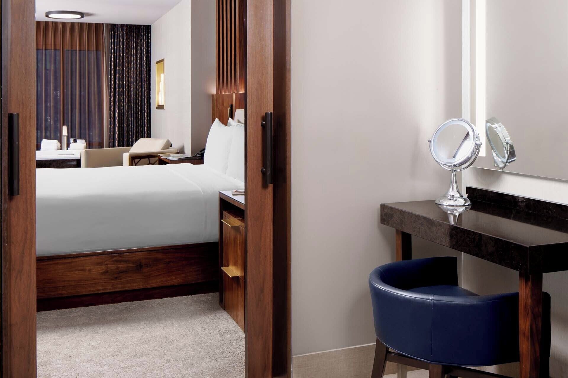 A hotel room with a bed, a desk with a mirror, a chair, and a window with curtains.