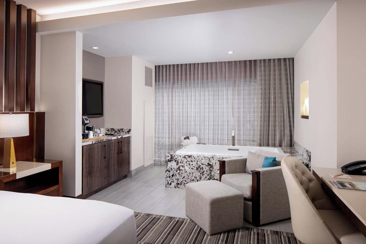 Une chambre d’hôtel comprenant un lit, un bureau, une chaise, une petite table, un canapé, une télévision et une salle de bain avec une baignoire et une douche.