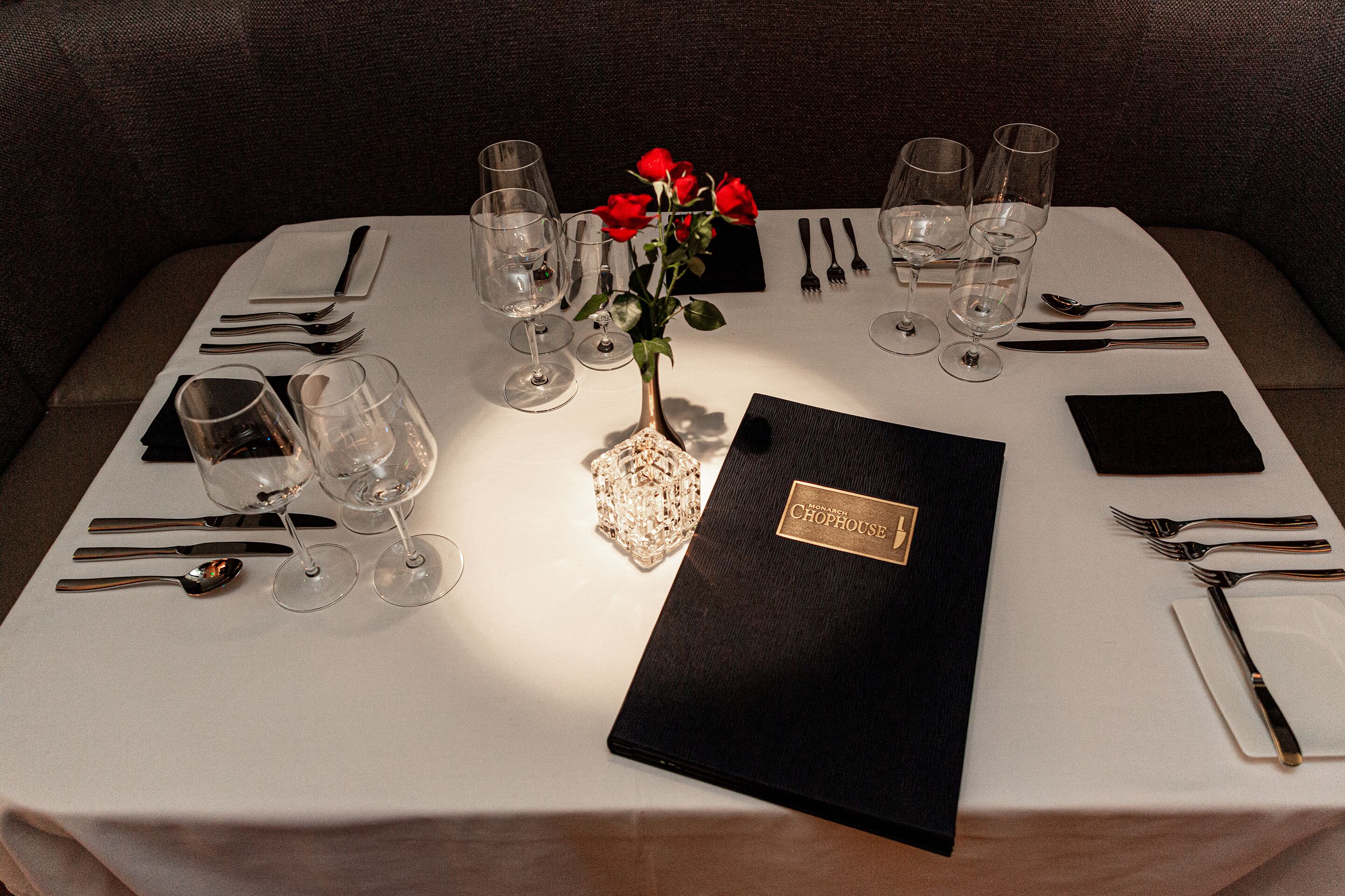 Una mesa lista para comer, con copas de vino, cubiertos y un florero con rosas rojas.