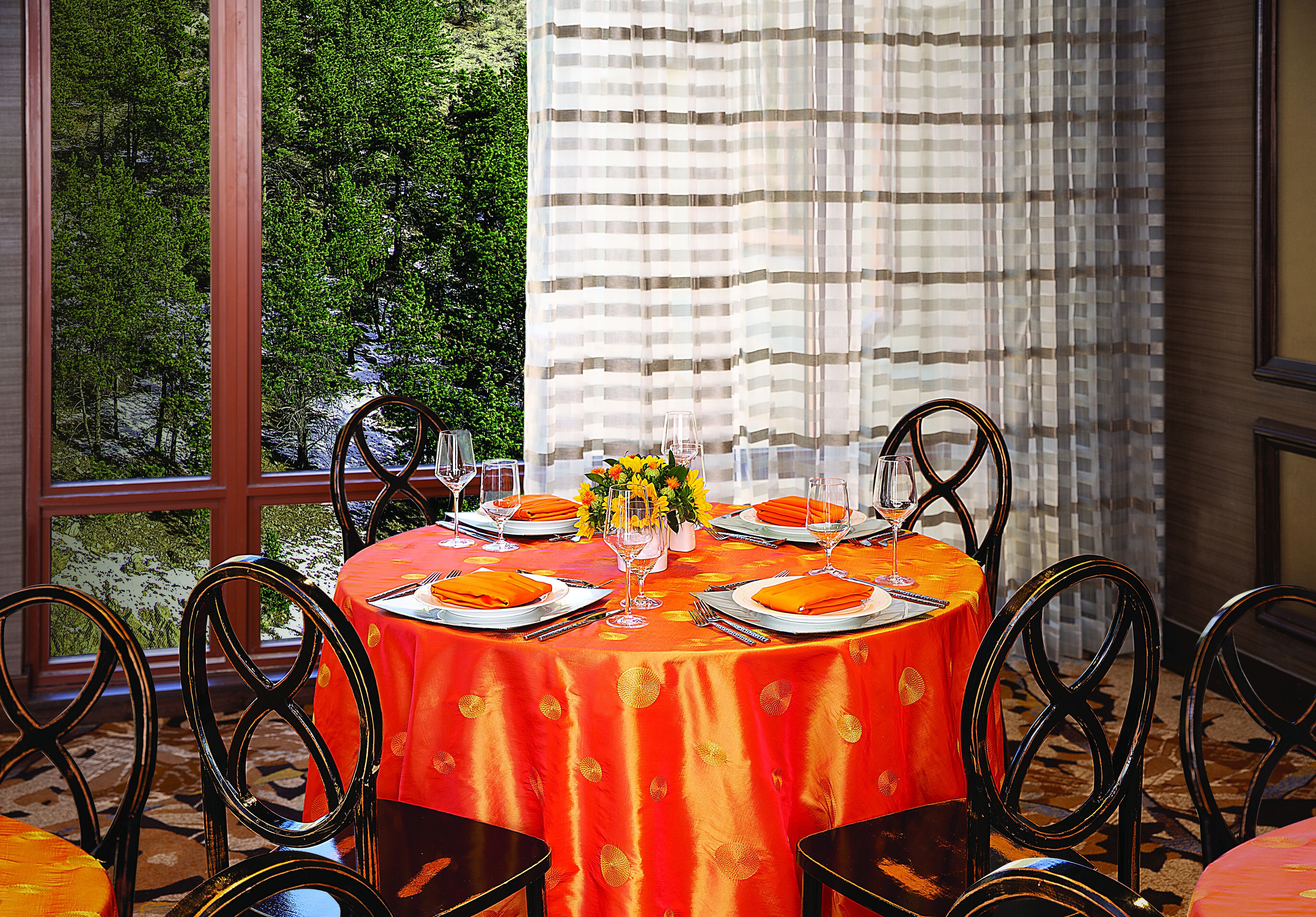 Mesa de comedor preparada para seis personas, con manteles naranjas, sillas negras y vistas a zonas verdes a través de amplios ventanales.