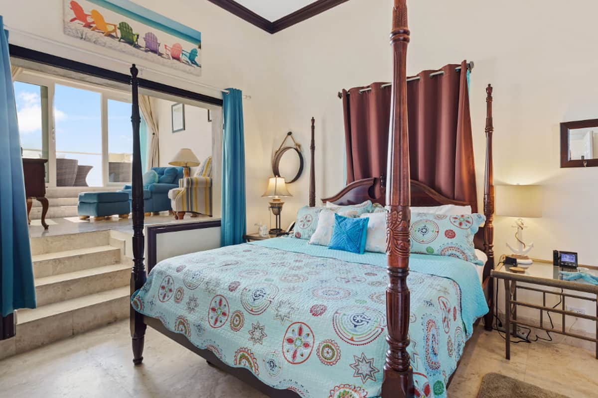 A four-poster bed with a patterned bedspread, a nightstand with a lamp, and a view of the outside through a glass door.