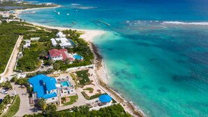 An aerial view of a coastal resort with a beach, clear turquoise waters, and a large blue-roofed building with a pool. - Villa Anguillitta (Blowing Point)