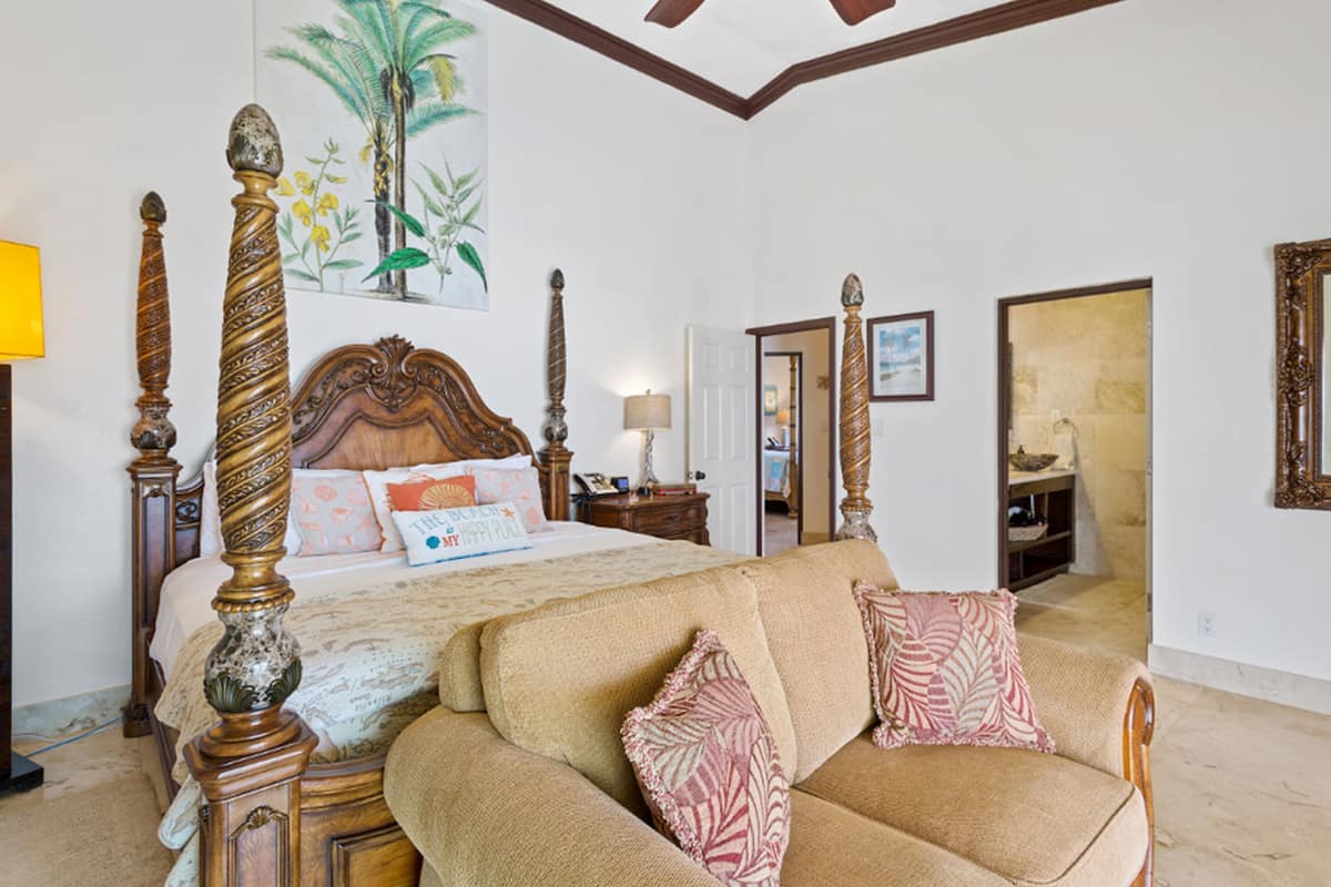 A bedroom with a large bed, a sofa, a painting of palm trees, and a wooden ceiling.