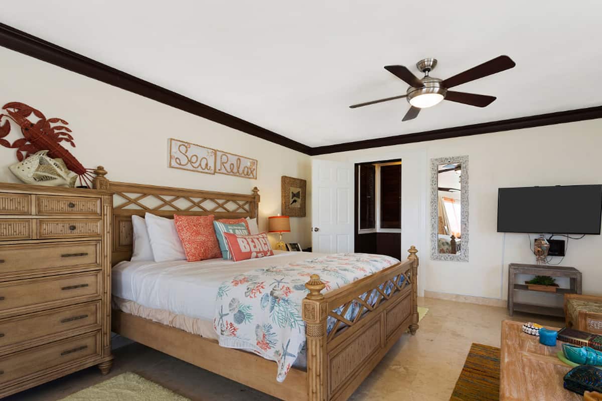 A bedroom with a bed, dresser, ceiling fan, and a TV on the wall.