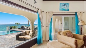 A luxurious room with a large window overlooking a pool and the sea, a comfortable sofa, and a dining area. - Villa Anguillitta (Blowing Point)