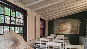 Een kamer met een eethoek, met witte stoelen en een tafel, een groot schilderij aan de muur, en uitzicht door een raam op groen buiten.