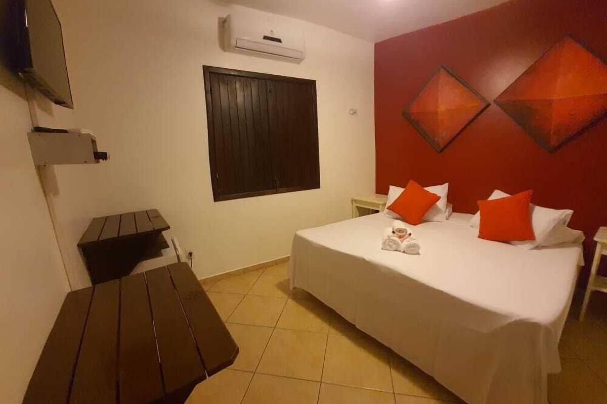 A hotel room with a bed, a TV mounted on the wall, a window, and a red accent wall.