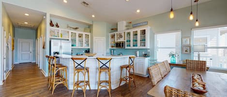 A spacious kitchen with a central island, wooden bar stools, and a dining area with a long table and wicker chairs.