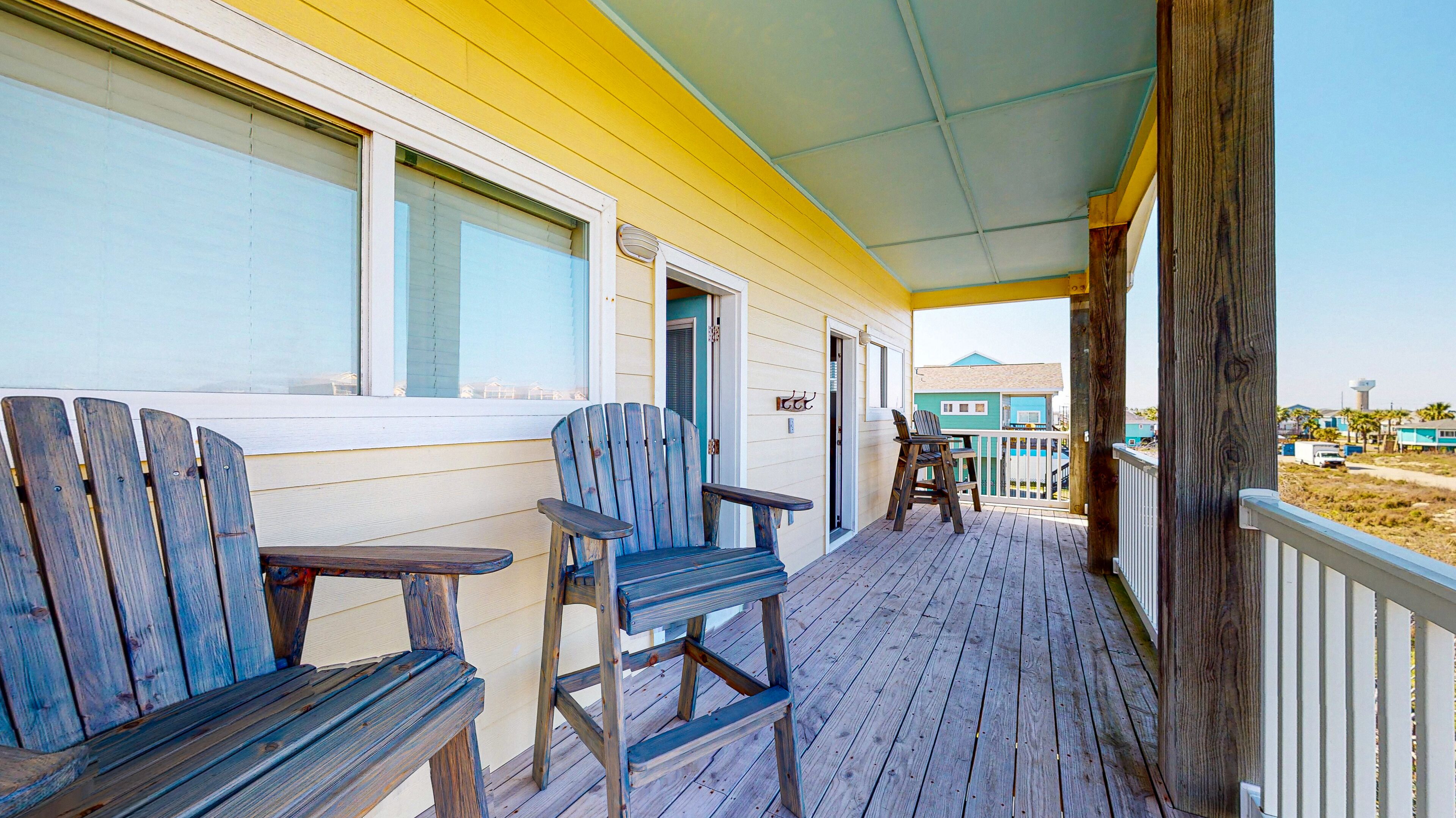 Une terrasse en bois avec deux chaises Adirondack et une petite table, donnant sur une plage où l’on aperçoit une maison bleue et un château d’eau au loin.