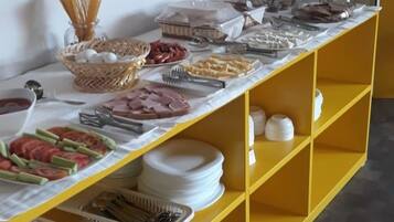Una mesa amarilla con varios alimentos y utensilios en una disposición de bufé frente a una pared lisa.