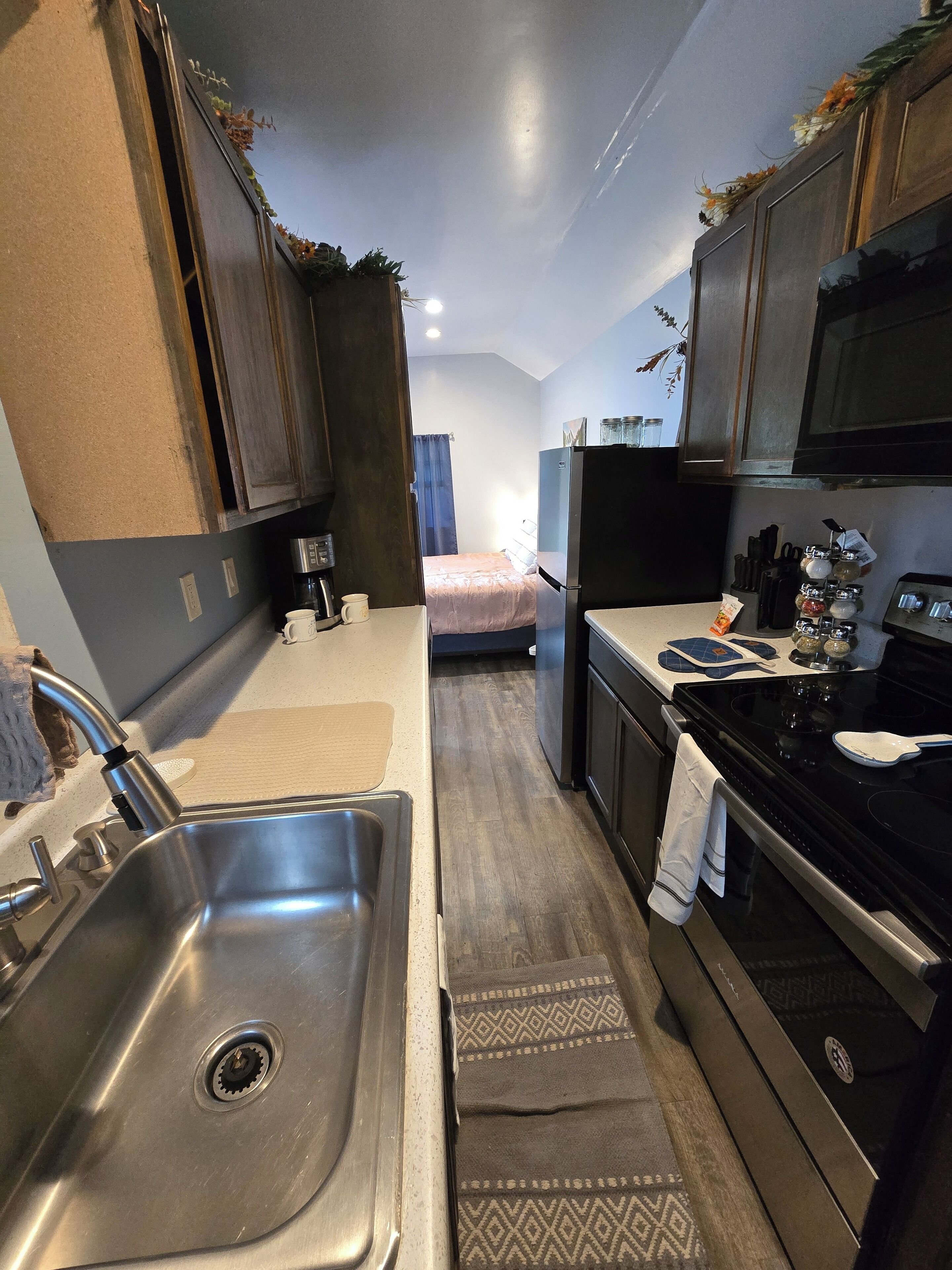 A compact kitchen with a sink, stove, and microwave. There is a bed in the background and a window with curtains.