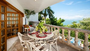 Un balcone con tavolo da pranzo apparecchiato per quattro, circondato da sedie bianche, con vista panoramica sul mare e sulla vegetazione lussureggiante.