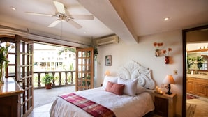 Una camera da letto con un letto grande, una ventola a soffitto, un balcone con piante e un bagno con specchio e lavandino.