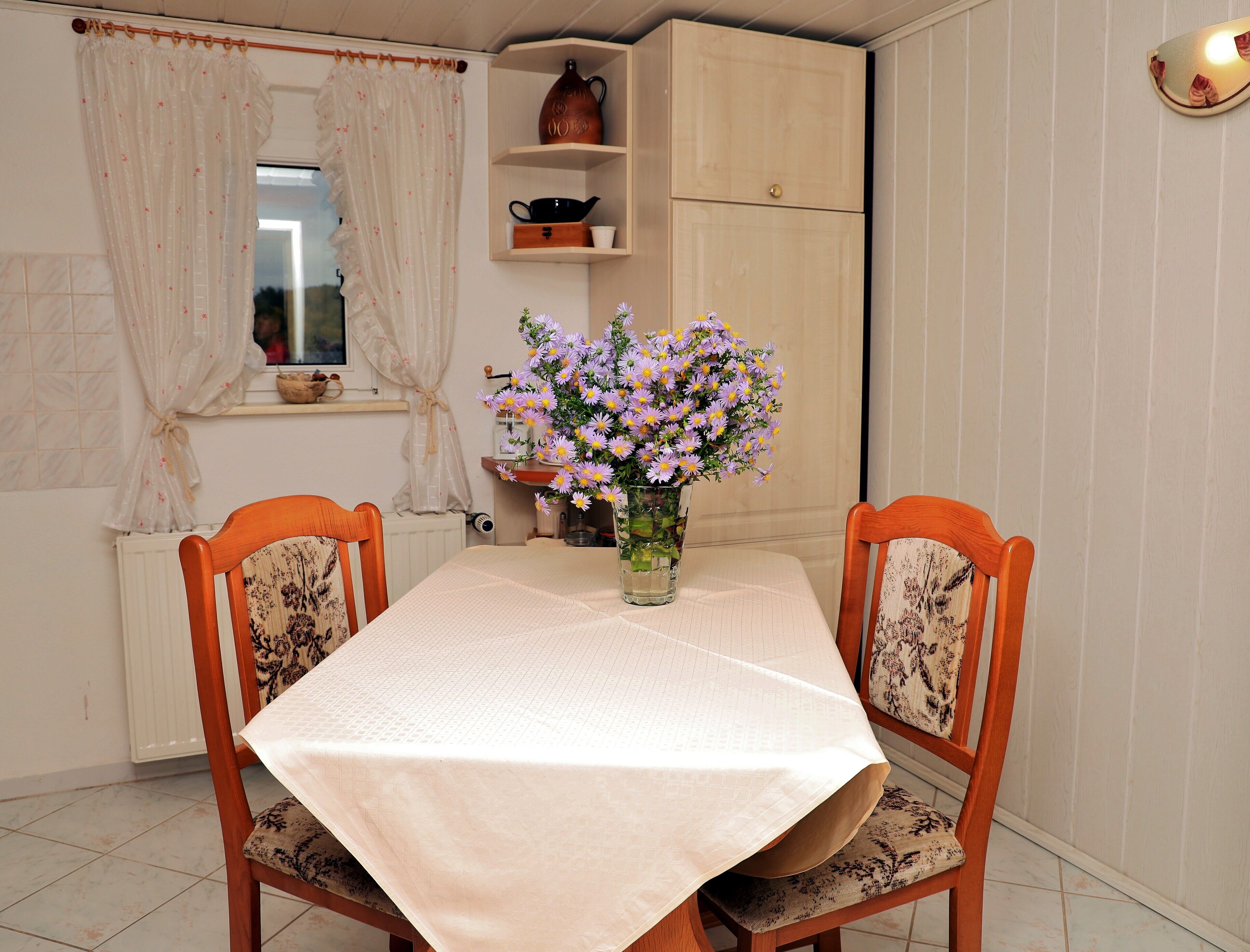 A dining area with a table set for two, a vase of flowers, and a cabinet above the table.