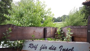 Un letrero de madera con el texto 'Reif für die Insel' en una tipografía rústica, sujeto a una valla de madera desgastada con follaje al fondo.