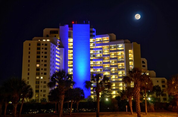 Un hôtel à plusieurs étages, doté de balcons illuminés et d’une tour centrale imposante, illuminée de lumière bleue.
