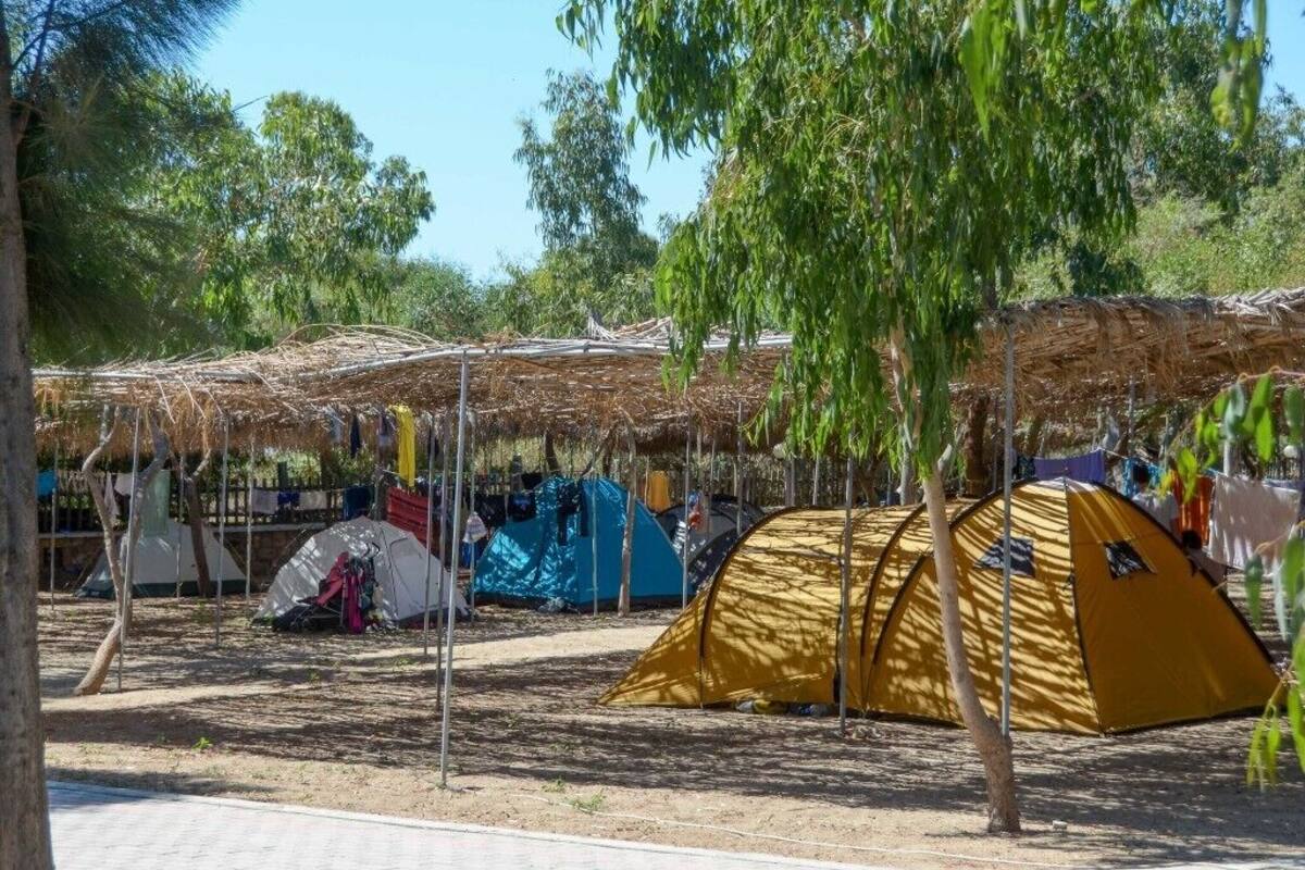 A campsite with several tents, including a prominent yellow tent, surrounded by trees and a thatched-roof structure.