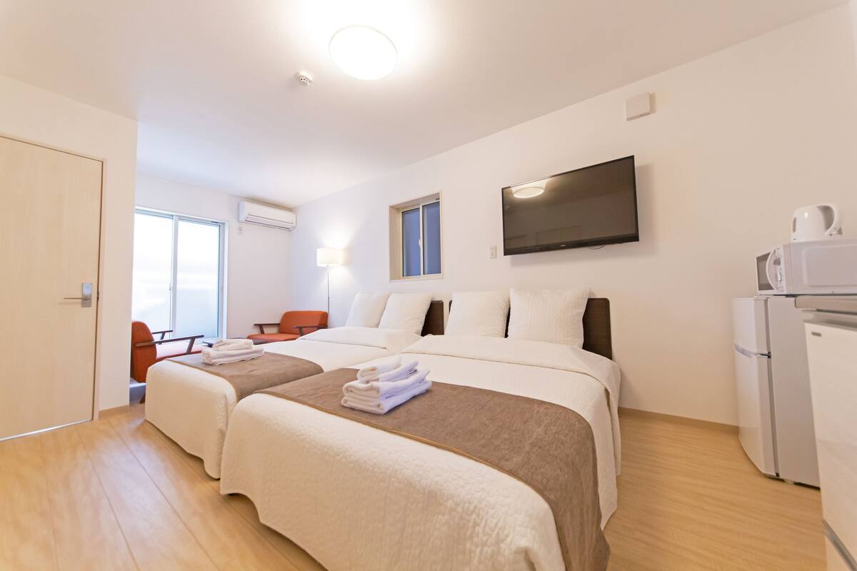A hotel room with two beds, a flat-screen TV, a refrigerator, and a window.