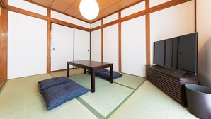 Ein traditioneller japanischer Raum mit Tatami-Böden, einem niedrigen Tisch, Kissen, einem Fernseher und einer Holzzieldecke.