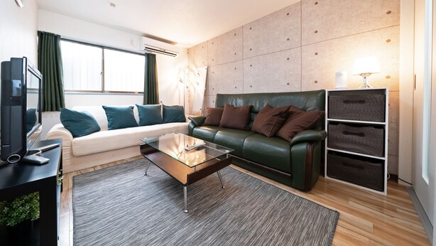 A modern living room with a glass coffee table, leather sofas, a flat-screen TV, and a large window with curtains. - TWS Morinomiya (Osaka)