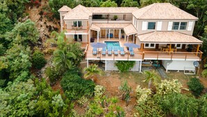 Una casa grande de varios pisos con piscina, rodeada de abundante vegetación.