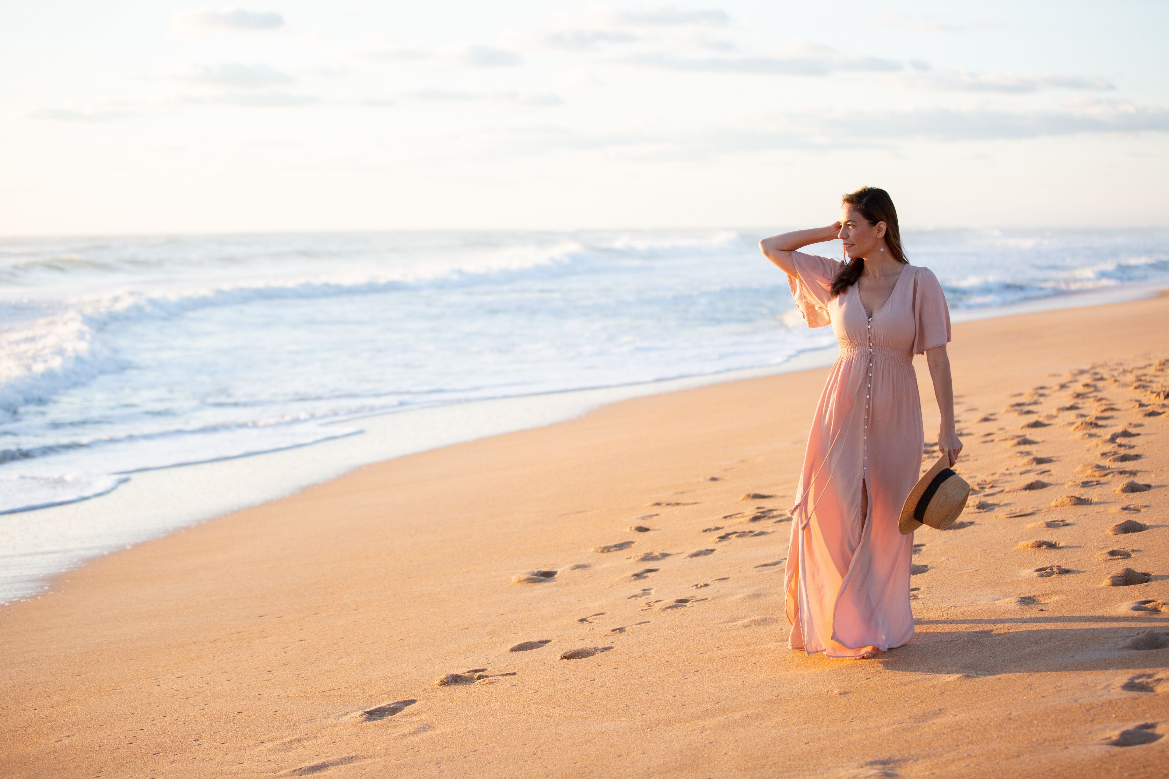 Eine Frau in einem langen Kleid steht auf einem Sandstrand, Fußabdrücke führen zum Meer.