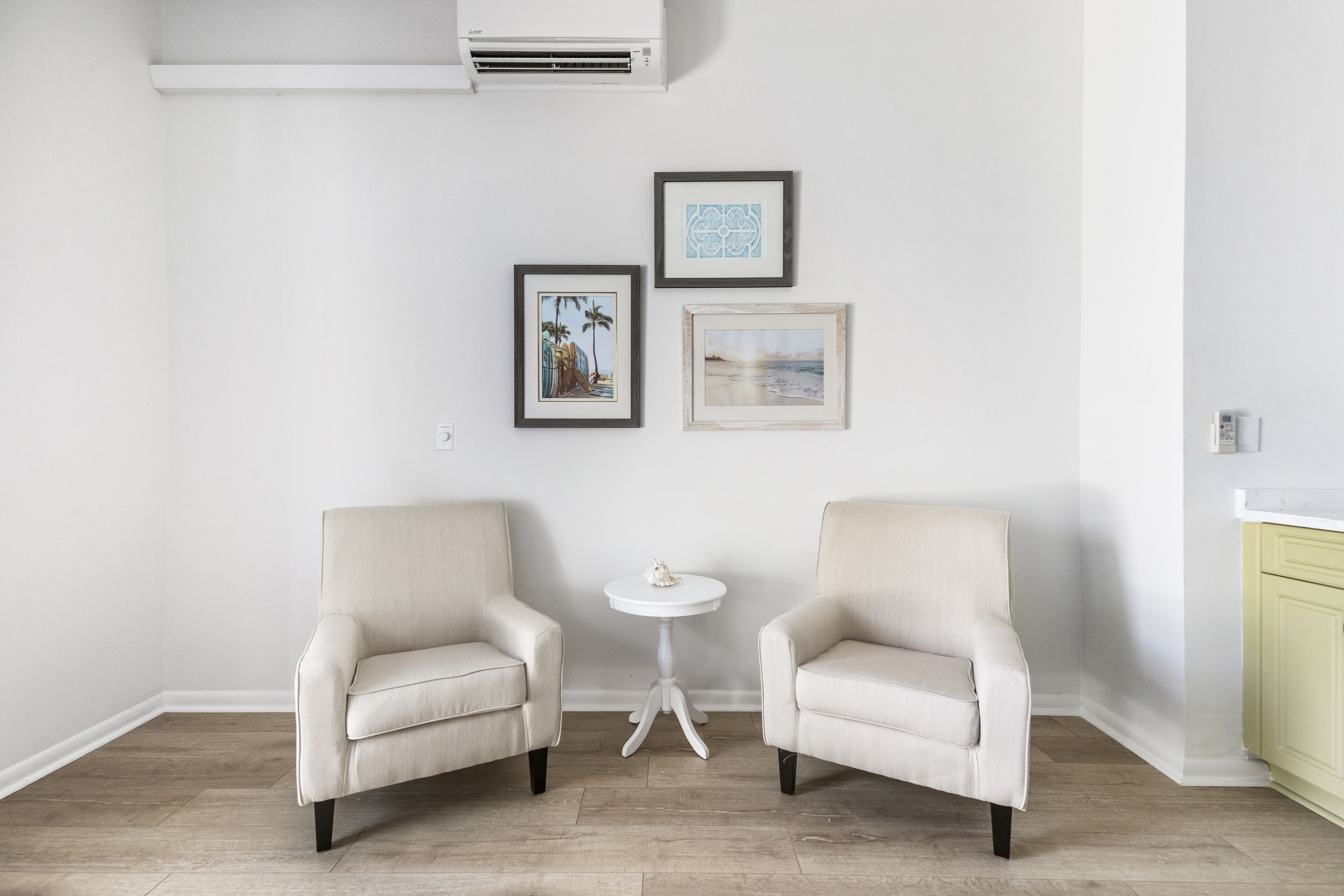 A room with two beige armchairs, a small white table, and three framed pictures on the wall.
