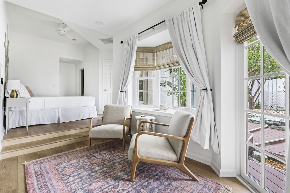 Ein Zimmer mit einem Bett, zwei Stühlen, einem kleinen Tisch, einem gemusterten Teppich und großen Fenstern mit Vorhängen.