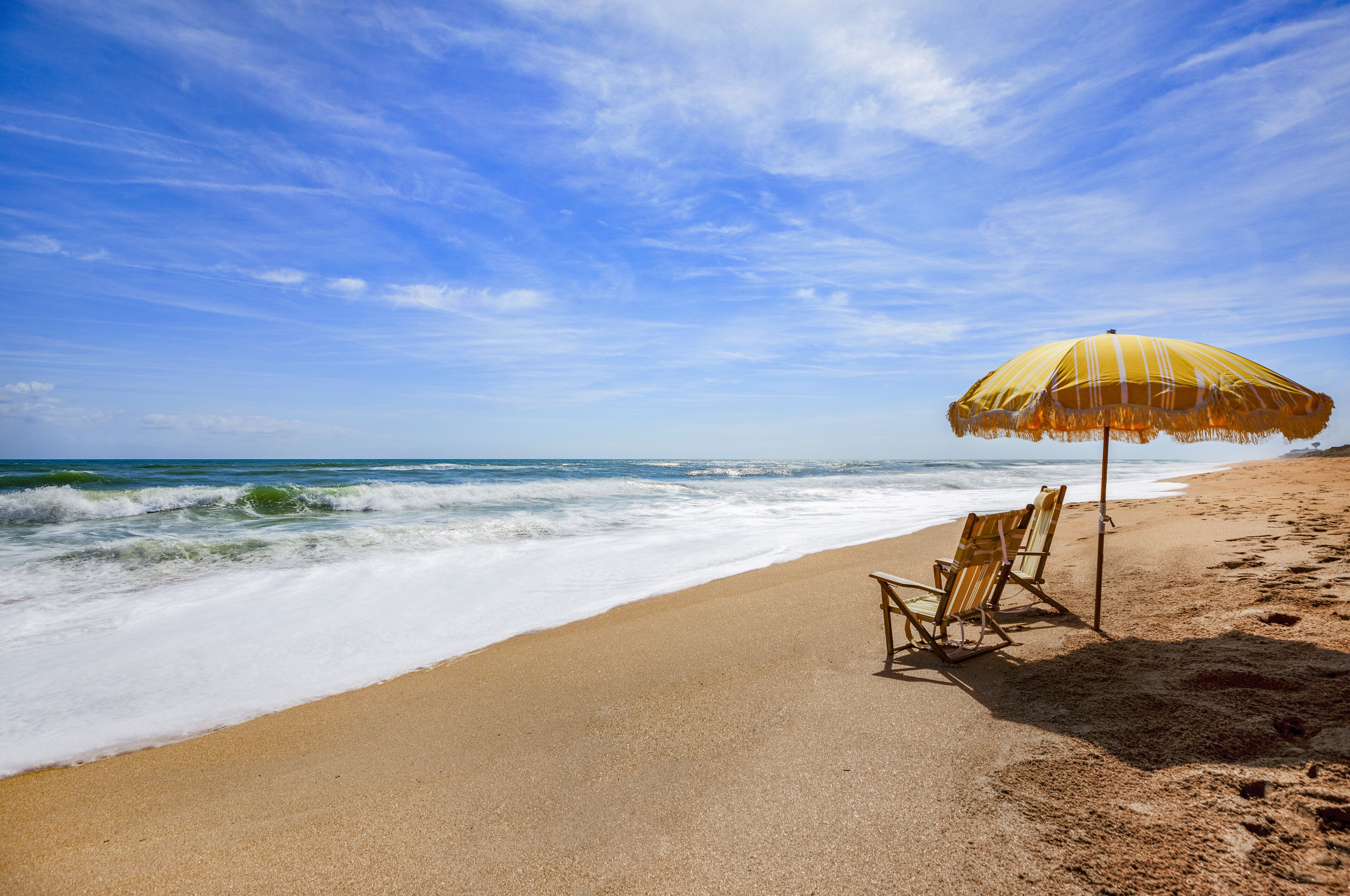 A beach with a wooden chair and a yellow striped umbrella.