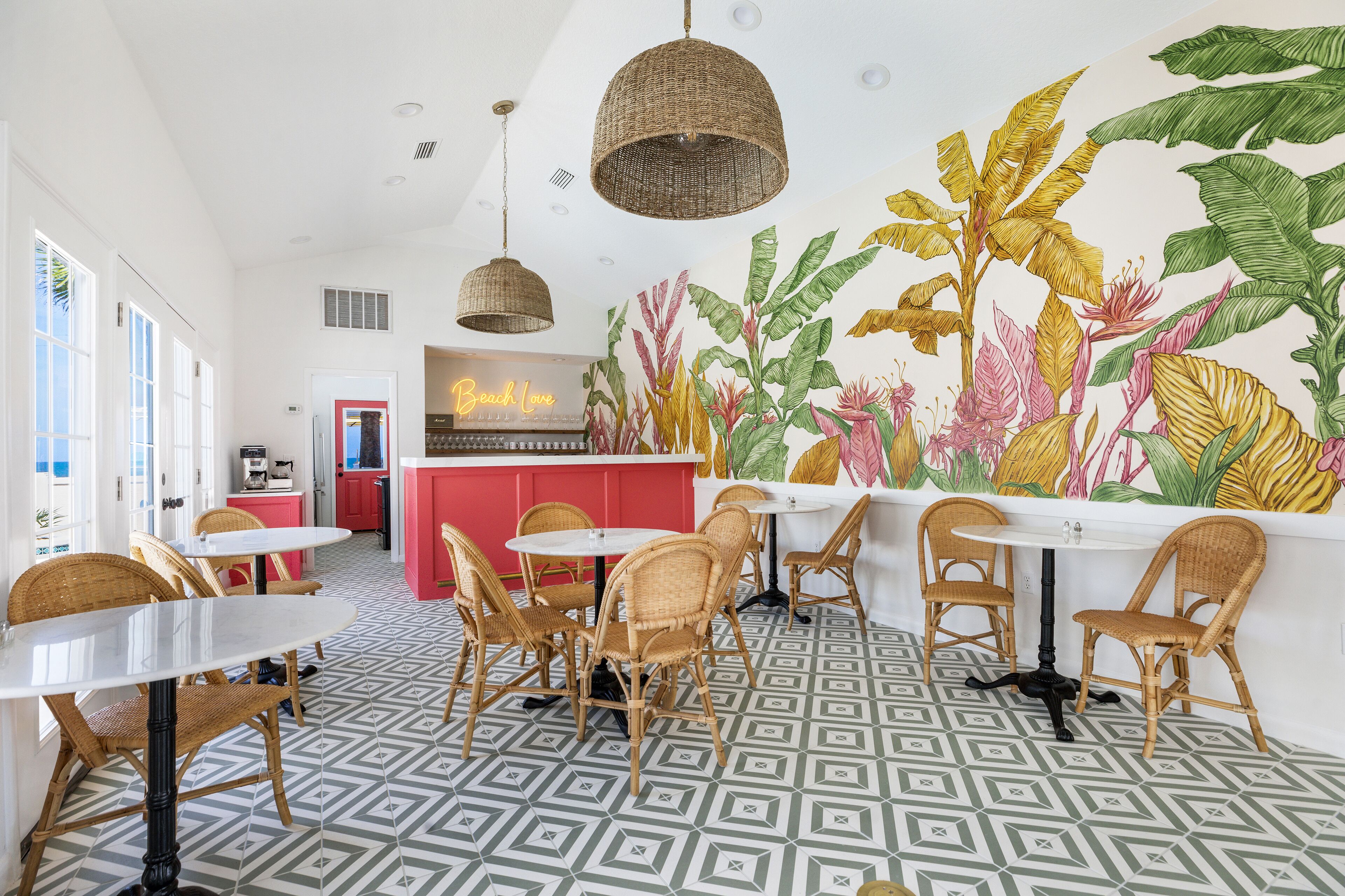 A restaurant with wicker chairs, white tables, and a mural of tropical plants.