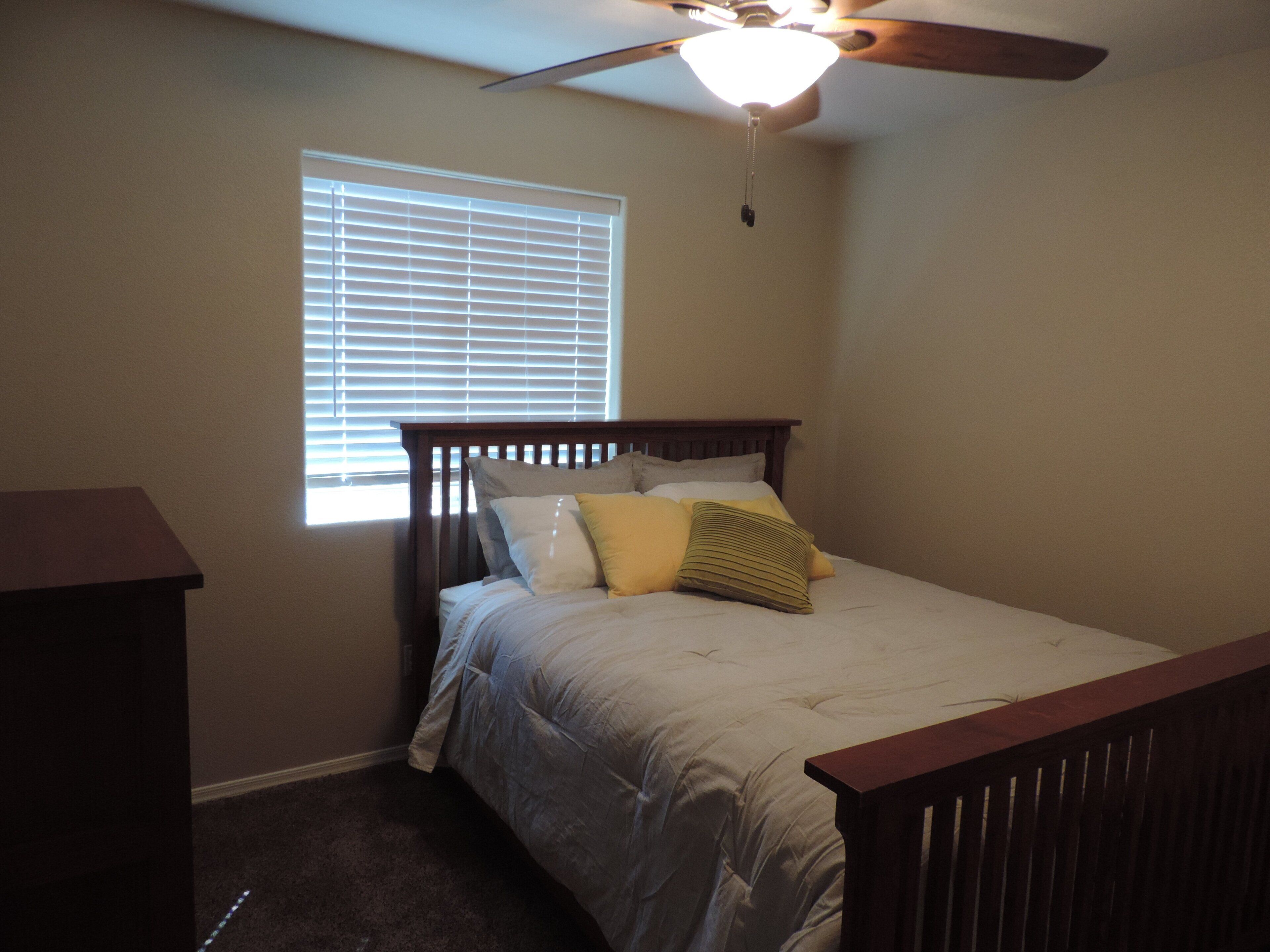 A bedroom with a bed, a ceiling fan, a window with blinds, and a wooden nightstand.