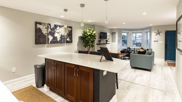A modern kitchen with a large island, wooden cabinets, and pendant lighting. There is a dining area with a gray sofa and a small table. - Lovely Shaw Apartment - Sleeps 4, WALK to White House (Washington)