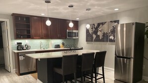 A modern kitchen with wooden cabinets, a stainless steel refrigerator, a microwave, and a bar area with stools. - Lovely Shaw Apartment - Sleeps 4, WALK to White House (Washington)