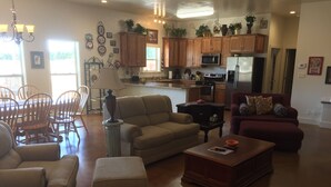 A spacious living room with a beige sofa, a red sofa, and a wooden coffee table. There is a dining area with wooden chairs and a chandelier. The kitchen features wooden cabinets, a microwave, and a refrigerator. - Willow Knoll 2 Texas (Fredericksburg)