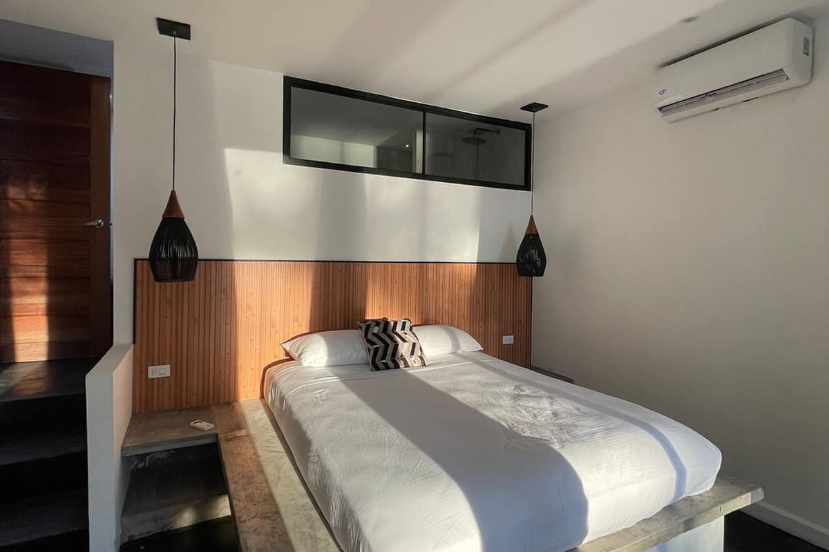 Une chambre moderne avec un grand lit, une tête de lit en bois, des luminaires suspendus et un appareil de climatisation.
